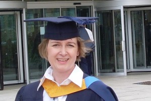 Picture Description: a woman wearing a mortarboard and gown, standing in front of some modern, institutional doors