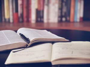 Picture description: Two books open on a table, one with a pen on it. A Shelf of books in the background.
