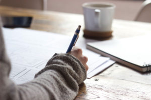 Picture Description: Someone's arm and hand resting on a desk, holding a pen and writing. They are wearing a grey woolly jumper. There is a mug of tea or coffee on the desk.